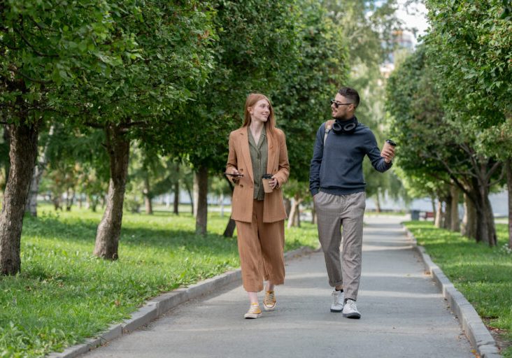 Two young colleagues in casualwear walking in large public park on summer day